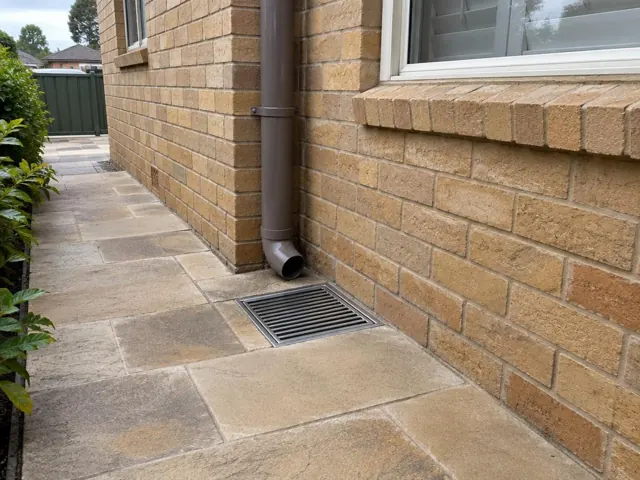 Outdoor gully grate beside a brick house wall and downpipe on dry paved path in Merrylands